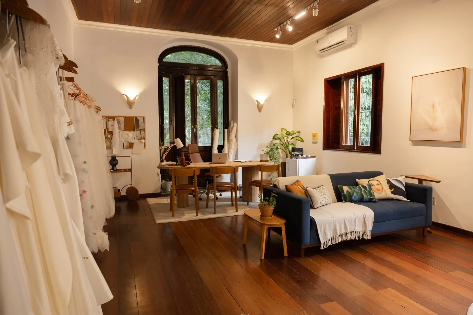 Sala de atendimento da Casa Belina com arara de vestidos de noiva, mesa de trabalho e sofá azul sob a janela em arco